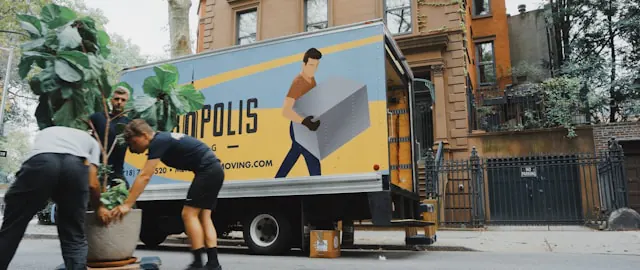 "Three movers are unloading a large potted plant from a moving truck parked on a city street. The truck features a bright yellow and blue design with an illustration of a man carrying a box and the company's name partially visible.